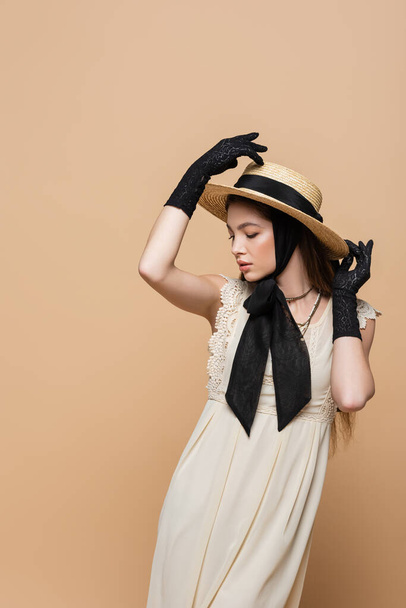 Stylish young woman in retro dress and gloves touching sun hat isolated on beige  - Фото, зображення
