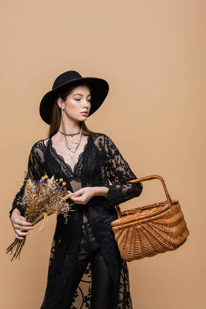 Stylish woman in fedora hat holding wicker basket and dry flowers isolated on beige  - Photo, image