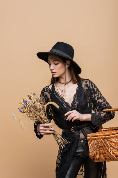 Young woman in guipure robe and fedora hat looking at dry flowers and holding basket isolated on beige  - Foto, Bild