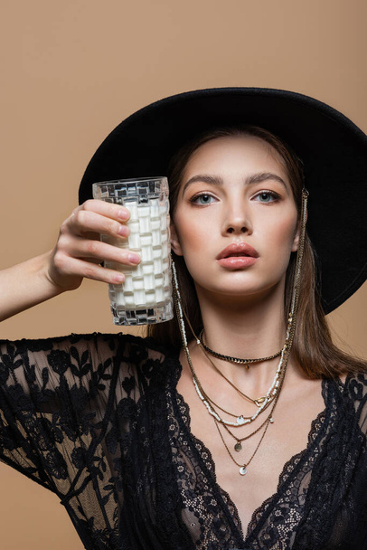 Portrait of fashionable model in fedora hat holding glass of milk isolated on beige  - Photo, Image