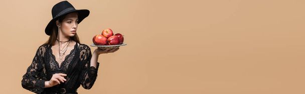Модная модель в шляпе fedora глядя на яблоки на тарелке изолированы на бежевый, баннер  - Фото, изображение