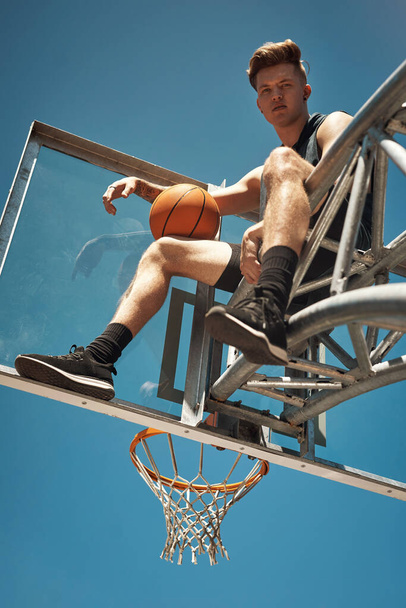 Πιάσε τον ουρανό ή μην προσπαθήσεις καν. Πορτρέτο ενός αθλητικού νεαρού που κάθεται σε στεφάνι μπάσκετ σε γήπεδο. - Φωτογραφία, εικόνα