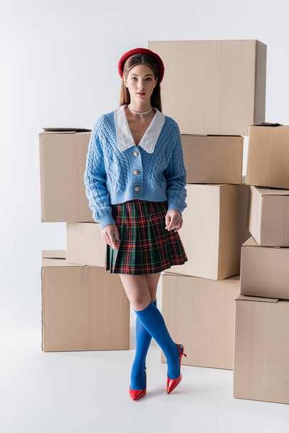Stylish woman in beret and skirt looking at camera near carton boxes on white background  - Photo, image