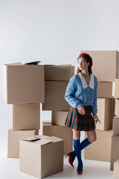 Young model in heels and beret standing near carton boxes on white background  - Fotó, kép