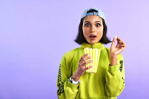 Если ты не знал, то теперь знаешь. Студийный снимок красивой молодой женщины, кушающей попкорн на фиолетовом фоне - Фото, изображение