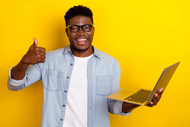 Photo of funky young brunet guy hold laptop thumb up wear eyewear jeans shirt isolated on yellow color background. - Valokuva, kuva