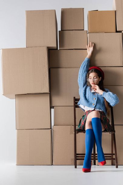 Fashionable woman in eyeglasses and heels sitting on chair near cardboard boxes on white background  - Photo, Image