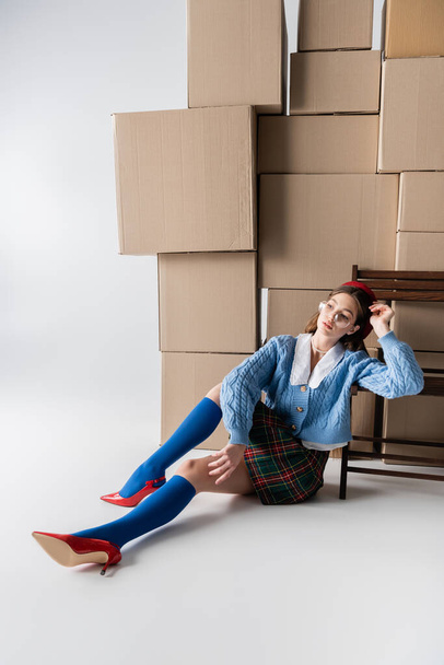 Trendy woman in eyeglasses and beret sitting near cardboard boxes and chair on white background  - Foto, afbeelding