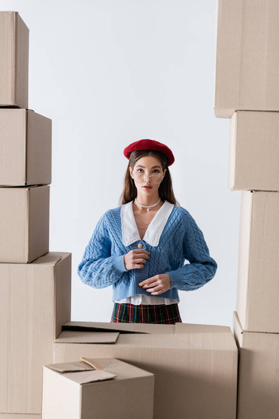 Pretty young woman in eyeglasses and beret touching knitted cardigan near cardboard boxes isolated on white  - Фото, зображення