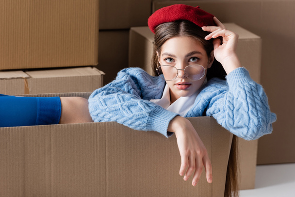 Woman in eyeglasses and cardigan sitting in package and looking at camera near cardboard boxes on white background  - Foto, afbeelding