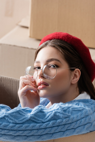 Portrait of trendy woman in beret and eyeglasses looking at camera near cardboard boxes  - Foto, Imagem