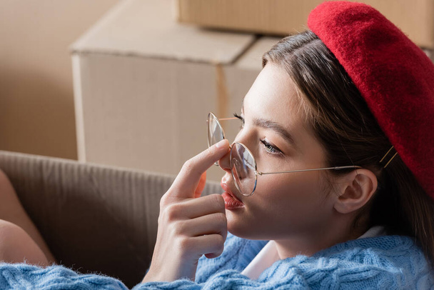 Young woman in beret touching eyeglasses near blurred cardboard boxes  - Фото, изображение