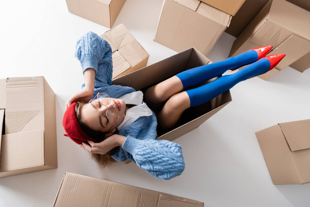 Top view of stylish woman in beret and eyeglasses sitting in package near cardboard boxes on white background  - 写真・画像