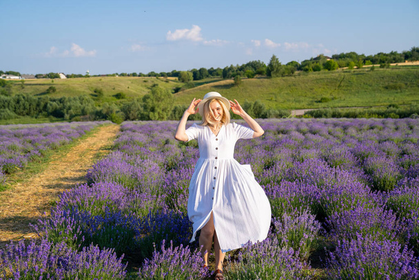 woman model in white dress outfit with hat is standing dancing in lavender field, photo session. Concept of freedom. Good picture for cover, calendar, postcard, wallpaper, background, product, website, blog, business, magazine, shop and store - Valokuva, kuva