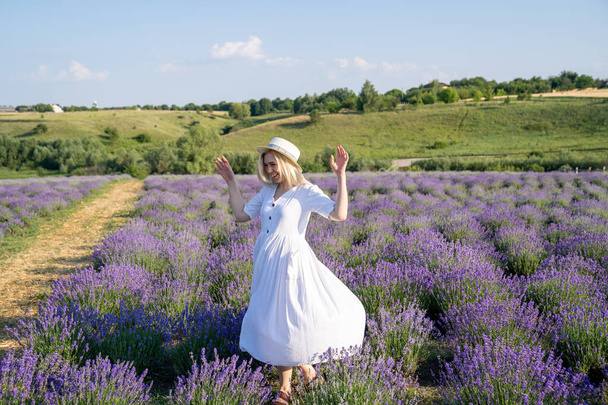 woman model in white dress outfit with hat is standing dancing in lavender field, photo session. Concept of freedom. Good picture for cover, calendar, postcard, wallpaper, background, product, website, blog, business, magazine, shop and store - Fotó, kép
