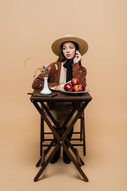 Trendy young woman in vintage clothes sitting near food and plants on table on beige  - Foto, immagini
