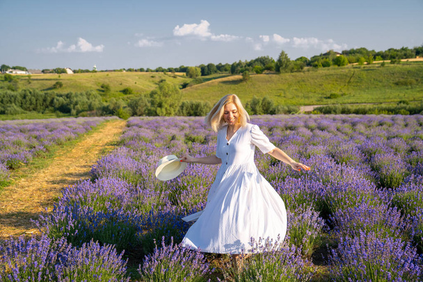 woman model in white dress outfit with hat is standing dancing in lavender field, photo session. Concept of freedom. Good picture for cover, calendar, postcard, wallpaper, background, product, website, blog, business, magazine, shop and store - Fotografie, Obrázek