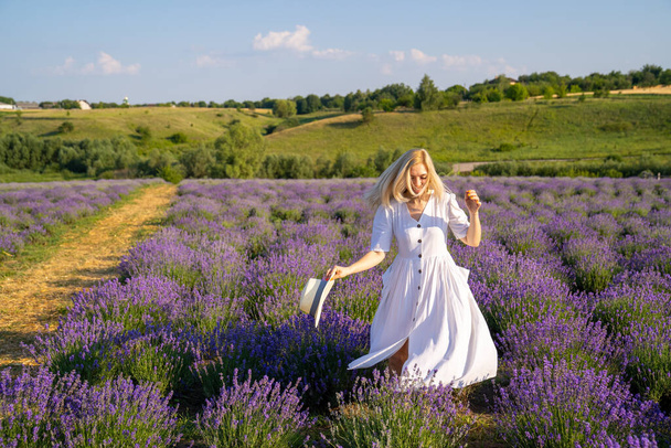 woman model in white dress outfit with hat is standing dancing in lavender field, photo session. Concept of freedom. Good picture for cover, calendar, postcard, wallpaper, background, product, website, blog, business, magazine, shop and store - Fotó, kép