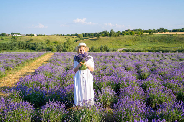 portrait of woman model in white dress outfit with hat is standing in lavender field, photo session. Young girl is holding lavender bouquet. Good picture for cover, calendar, postcard, wallpaper, background, product, website, blog, magazine, shop  - Foto, Imagem