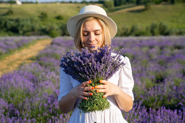 portrait of woman model in white dress outfit with hat is standing in lavender field, photo session. Young girl is holding lavender bouquet. Good picture for cover, calendar, postcard, wallpaper, background, product, website, blog, magazine, shop  - Fotó, kép
