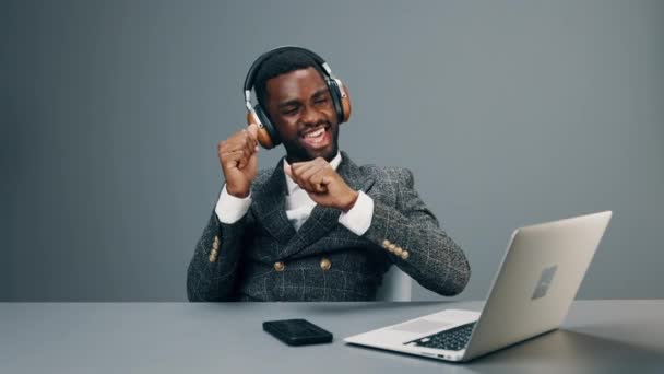 Een Afro-Amerikaanse zakenman zit in een koptelefoon aan zijn bureau en luistert naar vrolijke muziek terwijl hij danst en geniet van het leven op het werk. Hoge kwaliteit 4k beeldmateriaal - Video