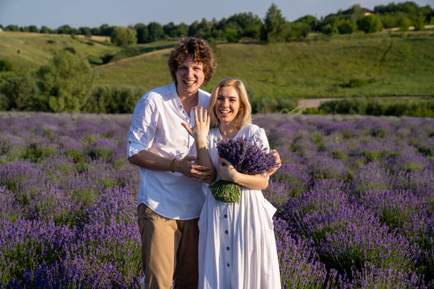 couple in white matching outfit in lavender field, photo session. man is proposing to woman with ring. engagement day. romance and true love in relationship. They are happy hugging and kissing. Engaged to get married  - Fotografie, Obrázek