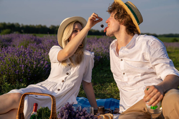 two persons couple in matching outfit have picnic in lavender field. They have fun, laugh and are feeding each other for photo session. Romantic relationship concept - 写真・画像
