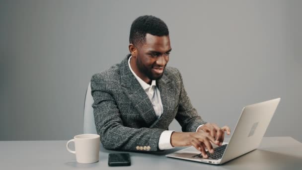 Uomo d'affari afroamericano in occhiali da computer lavora in ufficio dietro un computer portatile con una tazza di caffè alla sua scrivania su uno sfondo grigio ed è stanco. Filmati 4k di alta qualità - Filmati, video