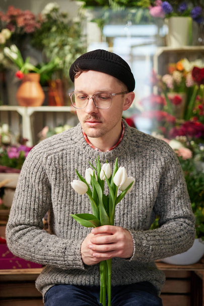 Attractive florist male in sweater and eyeglasses holding bouquet of white tulips. Fresh spring flowers. Mothers Day, Valentines Day or International Womens Day concept. High quality vertical photo - Zdjęcie, obraz