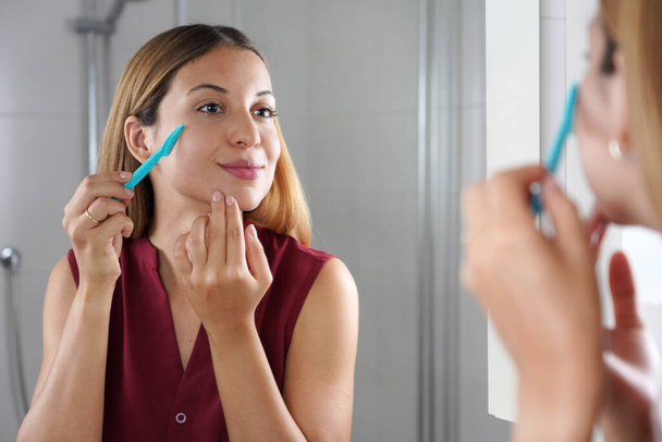 Facial hair removal. Brazilian girl shaving her face by razor at home. Beautiful young woman using razor on bathroom. - Fotoğraf, Görsel