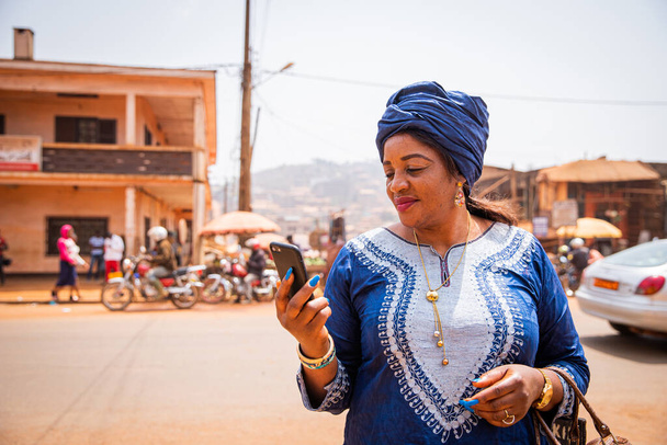 An African woman uses her smartphone while she is in the city center, she wears traditional african clothes - Photo, image