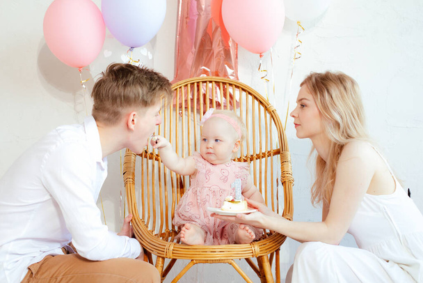 Delightful happy, festive full family of man and woman caring about small child feeding father by present cake sitting on chair indoors. Celebration event with balloons, one year old party holiday - Фото, зображення