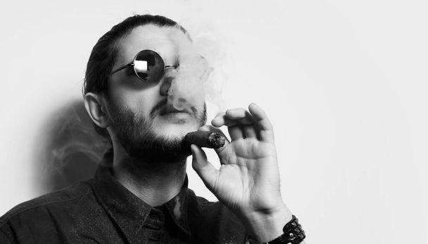 Черно-белый студийный портрет молодого красивого мужчины, курящего кубинскую сигару. - Фото, изображение