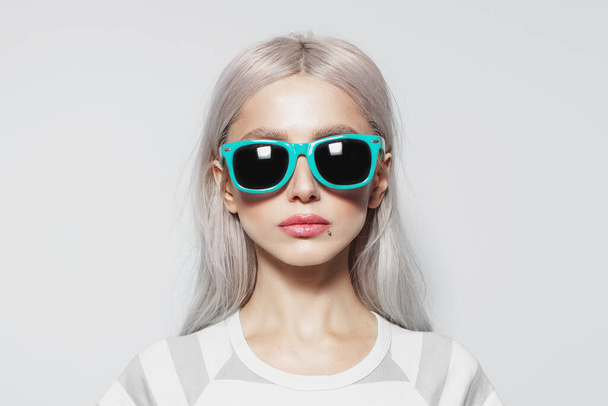 Studio portrait of serious, beautiful young girl with blonde hair in striped shirt on white background, wearing blue sunglasses. - Photo, Image