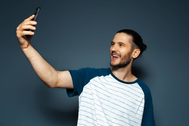 Studio portrait of young handsome smiling guy, taking selfie with smartphone. - Фото, изображение