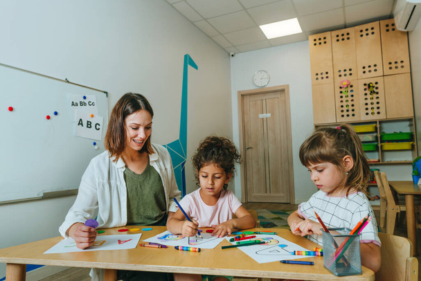 English lesson at elementary school or kindergarten. Students learning alphabet and colors with teacher coloring the letter A sitting in the classroom. Selective focus. - Fotografie, Obrázek