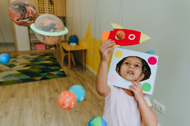 Funny little girl wearing handmade helmet playing with paper spaceship learning Solar system planets models at home or kindergarten. Education science concept. Selective focus. - Foto, imagen