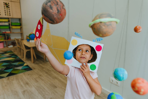 Funny little girl wearing handmade helmet playing with paper spaceship learning Solar system planets models at home or kindergarten. Education science concept. Selective focus. - Foto, afbeelding