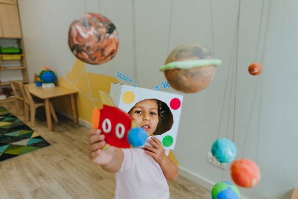 Funny little girl wearing handmade helmet playing with paper spaceship learning Solar system planets models at home or kindergarten. Education science concept. Selective focus. - 写真・画像