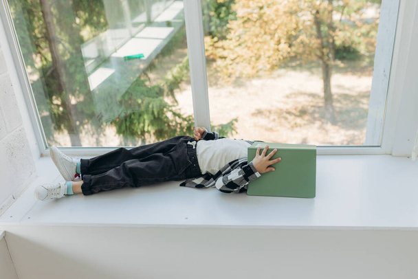 a tired schoolboy is lying on the windowsill reading a book. Learning at home - Fotografie, Obrázek