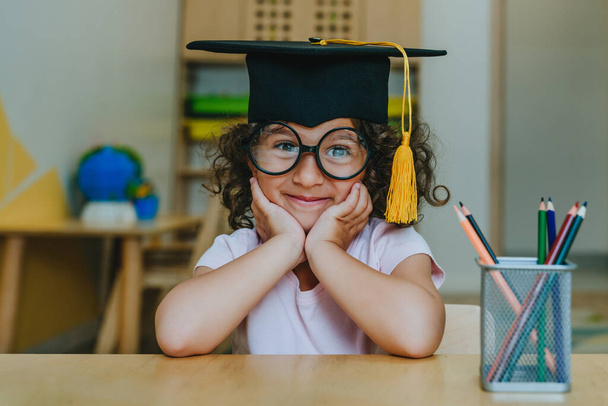 Portrait of happy little girl in graduation mortar hat sitting at desk at classroom - Photo, image