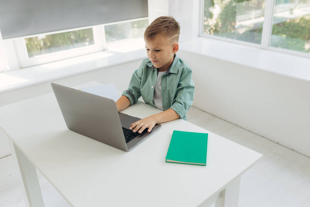 Αξιολάτρευτο εξυπηρετικό αγόρι γραπτώς στο σημειωματάριο και τη χρήση φορητού υπολογιστή, ενώ κάνει την εργασία. Θέση για κείμενο - Φωτογραφία, εικόνα