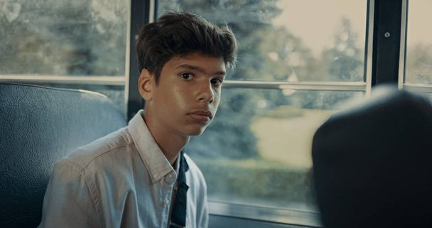 Pensif garçon indien aux cheveux bruns assis à la fenêtre de l'autobus scolaire seul fermer. Portrait de l'adolescent écolier effrayé tendu dans les transports étudiants. Élève mignon sérieux regardant la caméra attentivement. - Photo, image