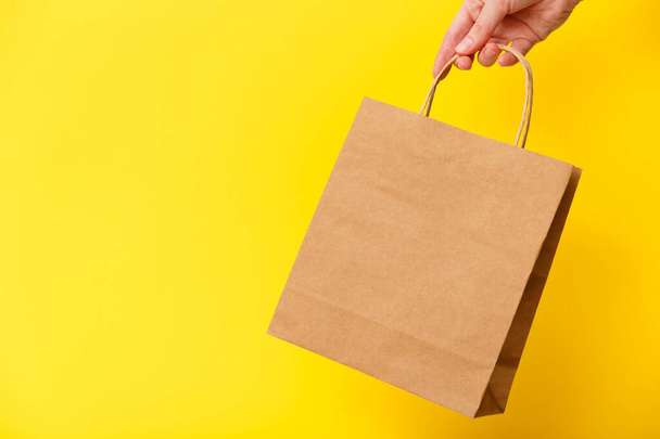 Mano femenina sosteniendo la bolsa de compras en blanco artesanal marrón aislada sobre fondo amarillo. Viernes negro venta, descuento, reciclaje, compras y ecología concepto - Foto, Imagen