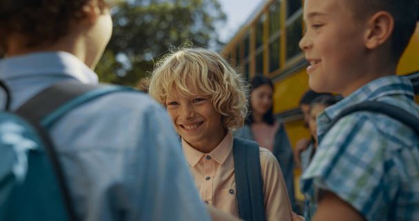 Drie scholieren lachen om de gele bus. Basisleeftijd jongens wachten op schoolbus boarding close-up. Leuke glimlachende leerlingen met rugzakken die samen plezier hebben in de buurt van het voertuig. Onderwijsconcept. - Foto, afbeelding