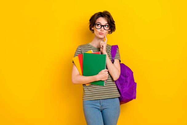 Photo of doubtful unsure lady wear striped t-shirt spectacles backpack looking empty space holding copybooks isolated yellow color background. - Фото, зображення