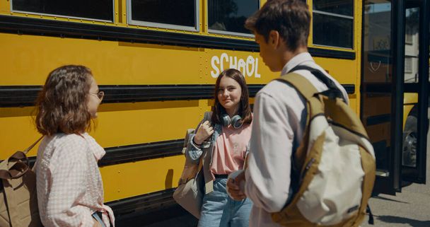 Cheerful teen classmates chatting at classic school bus. Three school children standing at yellow academic shuttle waiting boarding. Cheerful teenagers friends communicating outdoors before studies. - Foto, Imagen