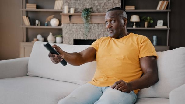 Elderly single bachelor african american adult senior man sitting on couch watching TV enjoy serial movie comments news program on television use remote control switch sport channels relaxing at home - Foto, Imagem
