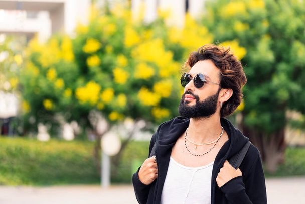 portrait of a stylish young college student with beard and sunglasses walking happy by the university campus, concept of youth and urban life style, copy space for text - Foto, Bild