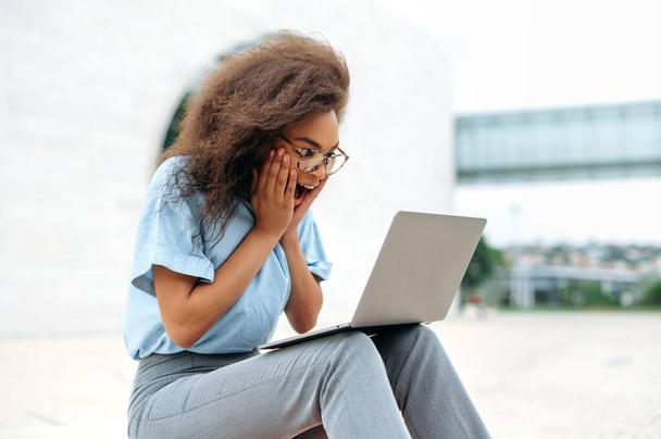 Shocked amazed stylish african american young woman, company worker, student, sitting with a laptop outdoors, looking at the screen in surprise, received unexpected news, rejoices, smiles - Photo, image